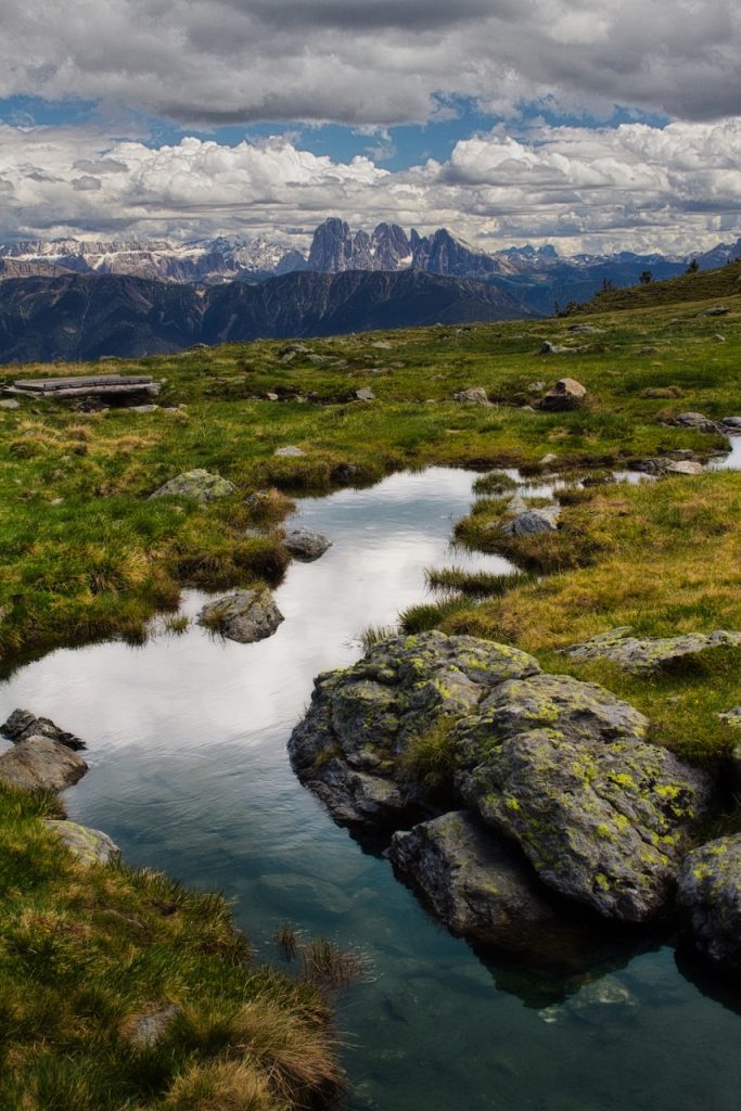 Pozze d'acqua specchiano il cielo tra rocce lichenate e cime dolomitiche all'orizzonte.
