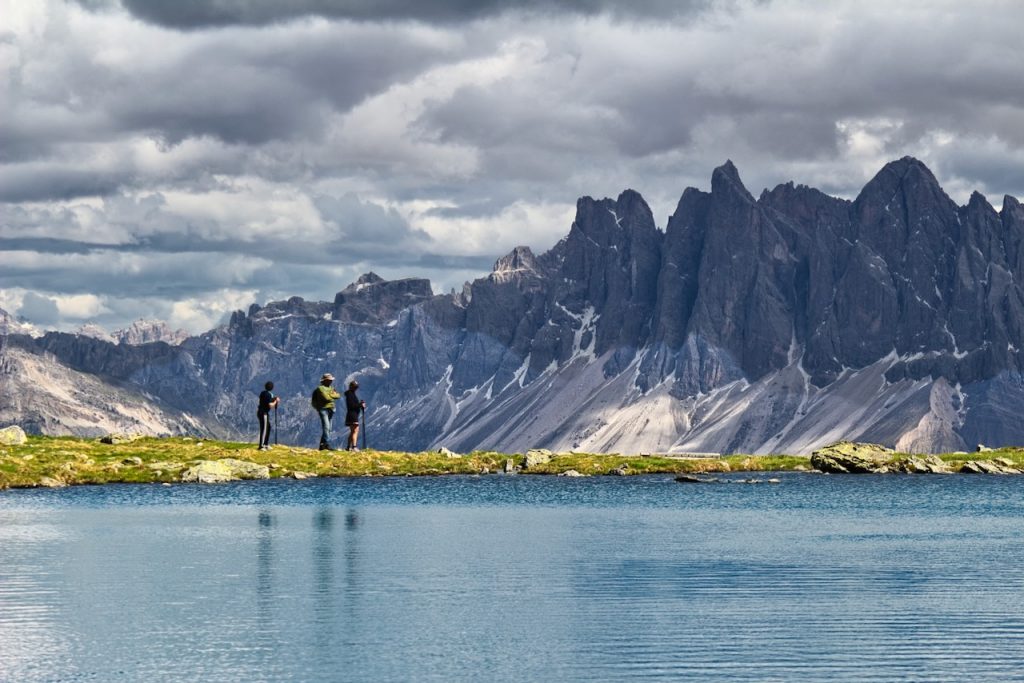 Tre escursionisti osservano le Odle, riflesse in un lago calmo e turchese.
