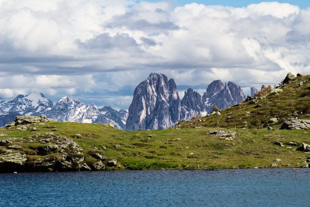 Il Sassolungo svetta oltre un lago blu, tra prati e cielo carico di nuvole.