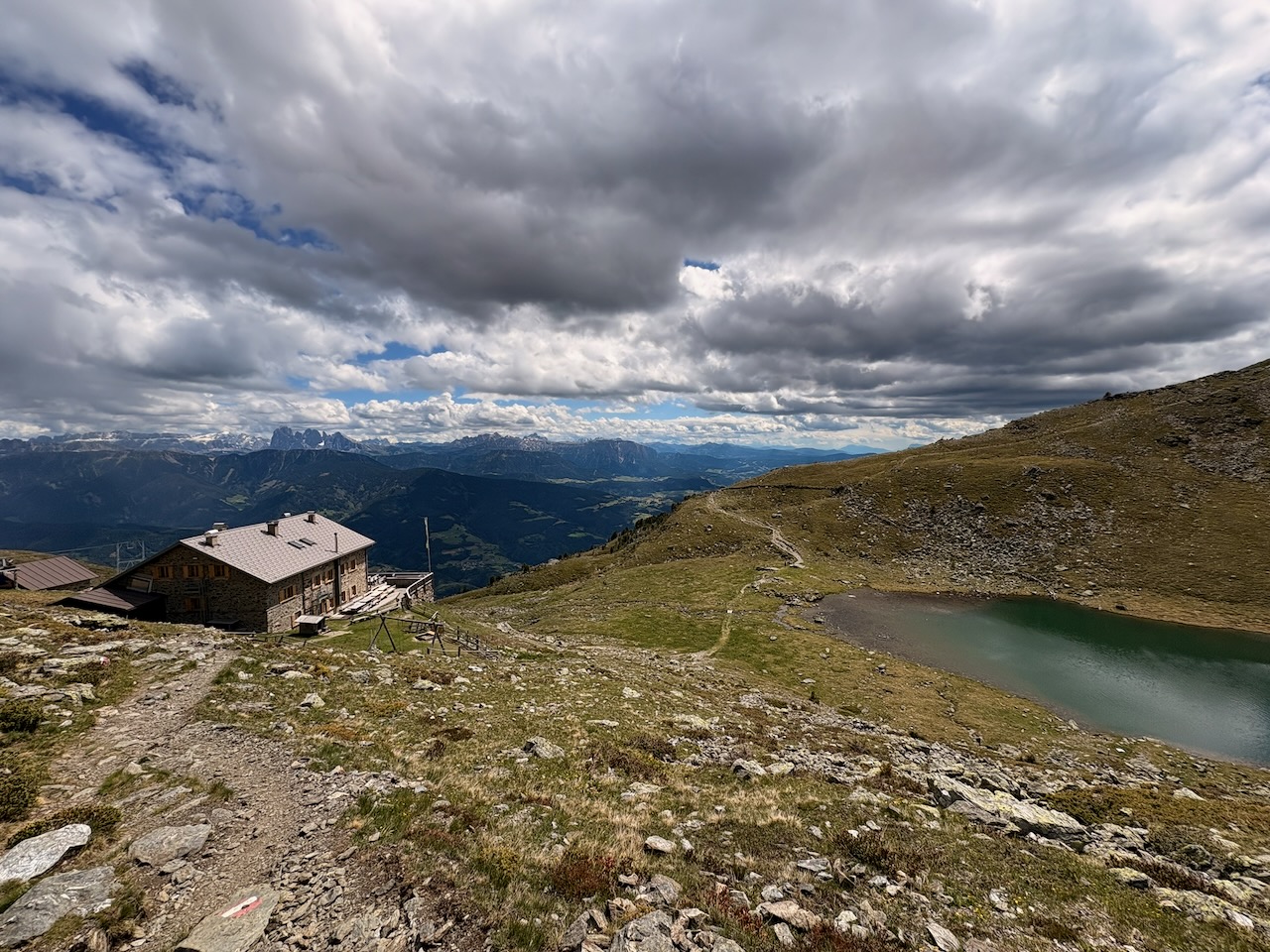 Rifugio di pietra affacciato su una valle profonda, accanto a un piccolo lago verde-azzurro.
