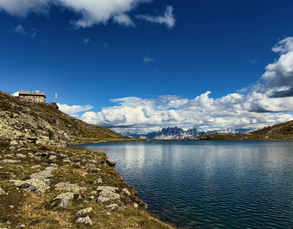 Lago alpino dalle acque blu intenso, circondato da rocce e licheni. Sulla sponda un rifugio in pietra domina il pendio, mentre in lontananza svettano le cime dolomitiche delle Odle, tra nuvole bianche e cielo terso.